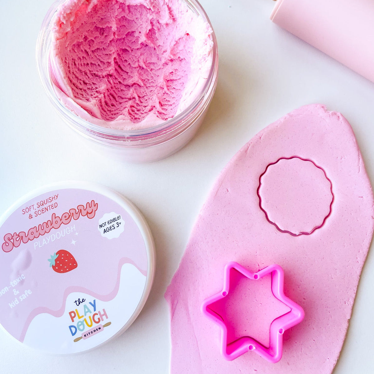 A jar of pink strawberry play dough on a white surface with cookie cutters and a rolling pin