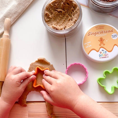 Child playing with gingerbread-themed play dough and tools on a white surface.