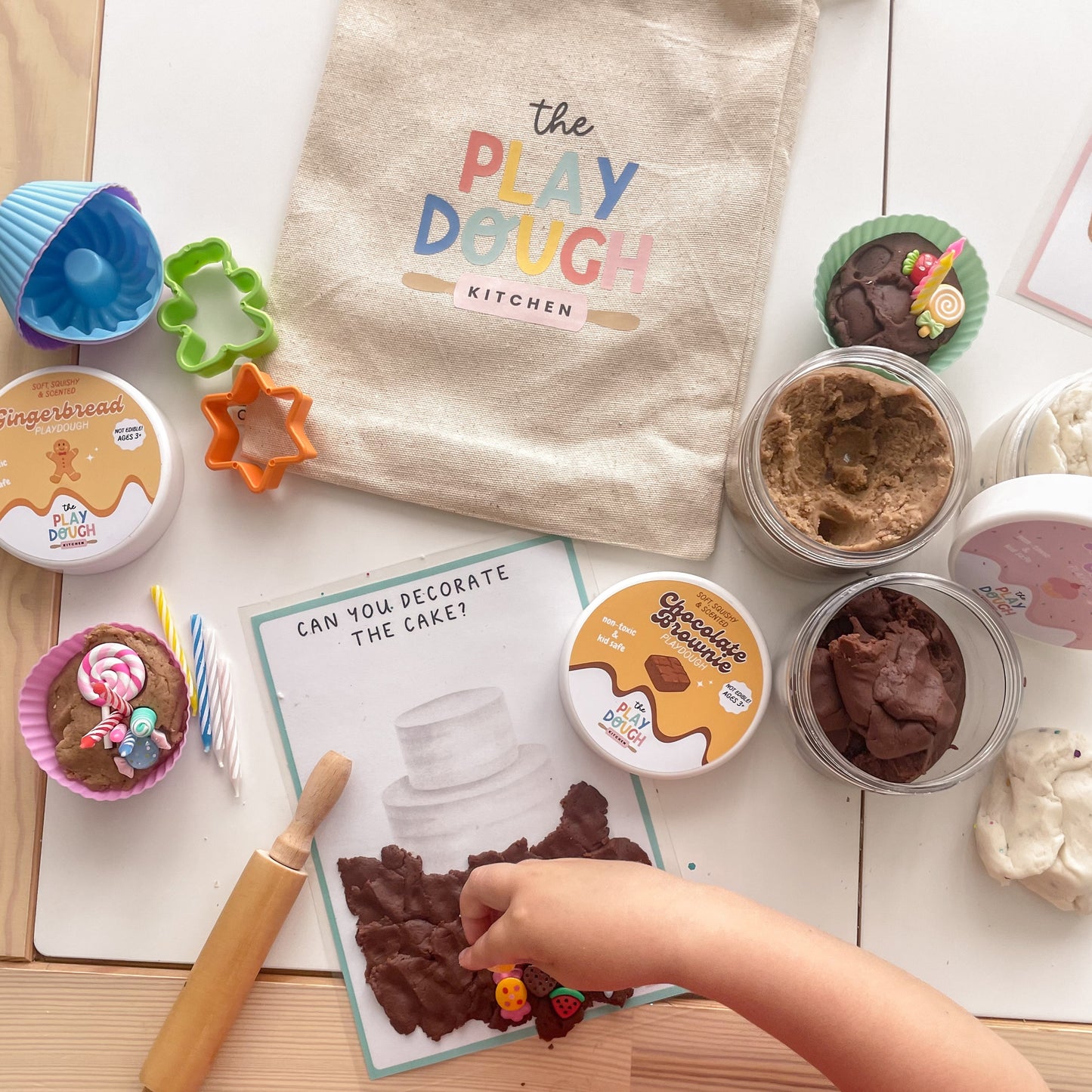 Baking themed play dough kit with containers, tools, and a child's hand interacting with the materials on a table.