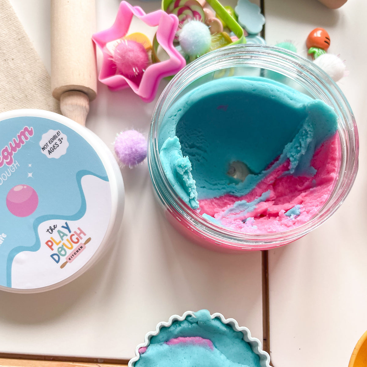 Play dough container with blue and pink bubblegum play dough, surrounded by tools and toys on a light surface.