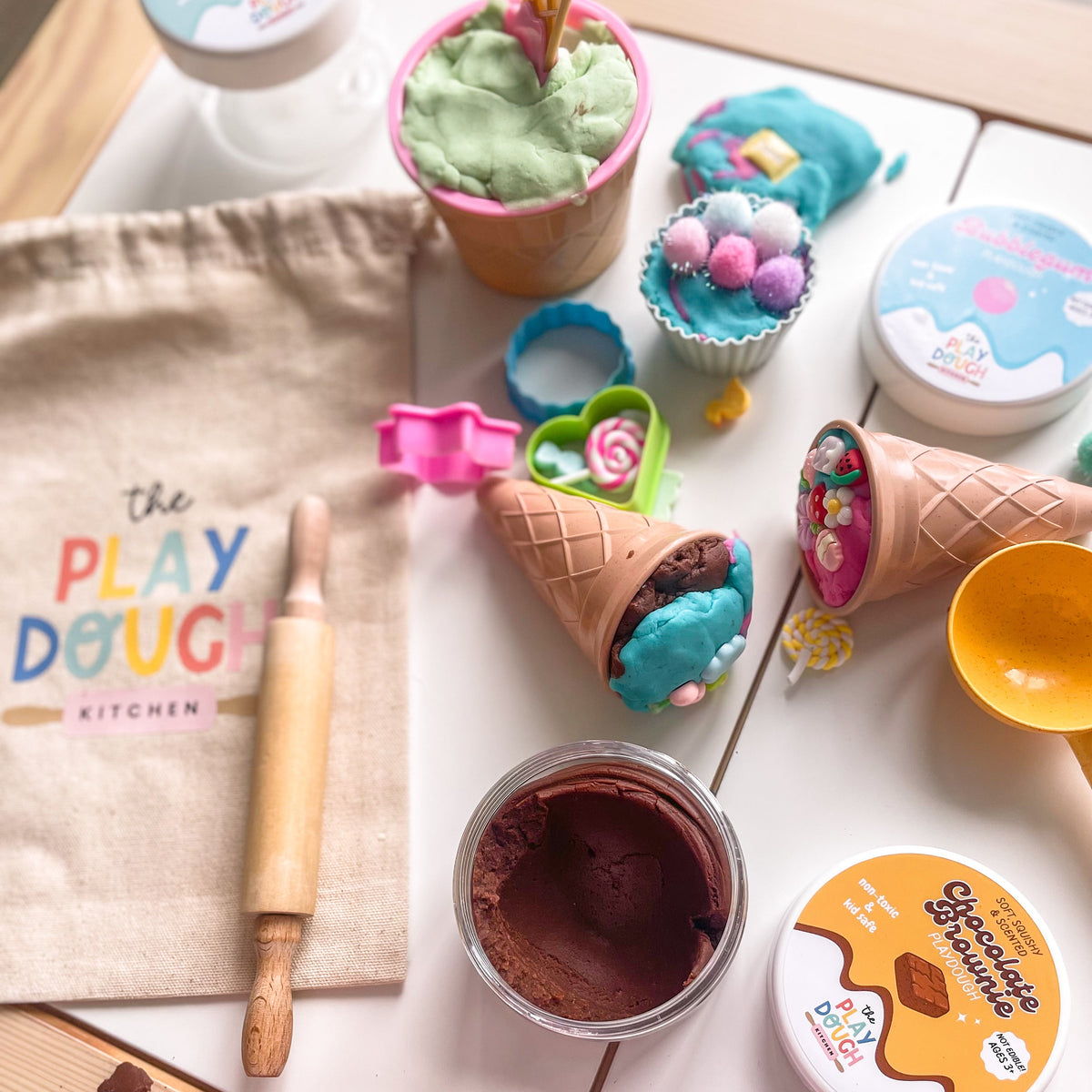Ice cream themed play dough kit with various play dough containers, tools, and a child's hands interacting with the items on a wooden surface.