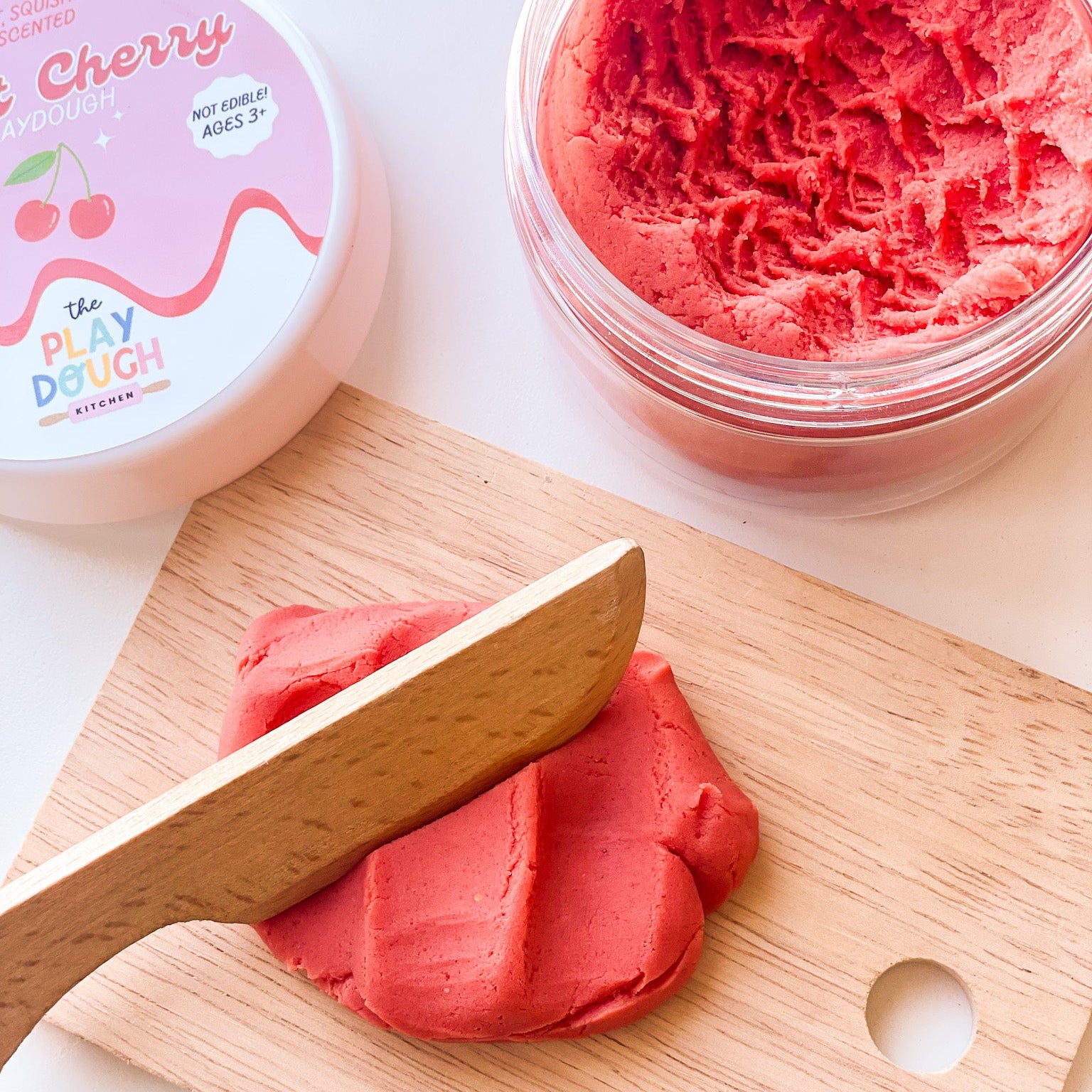 A jar of red playdough on a white surface with a kids wooden chopping board and knife