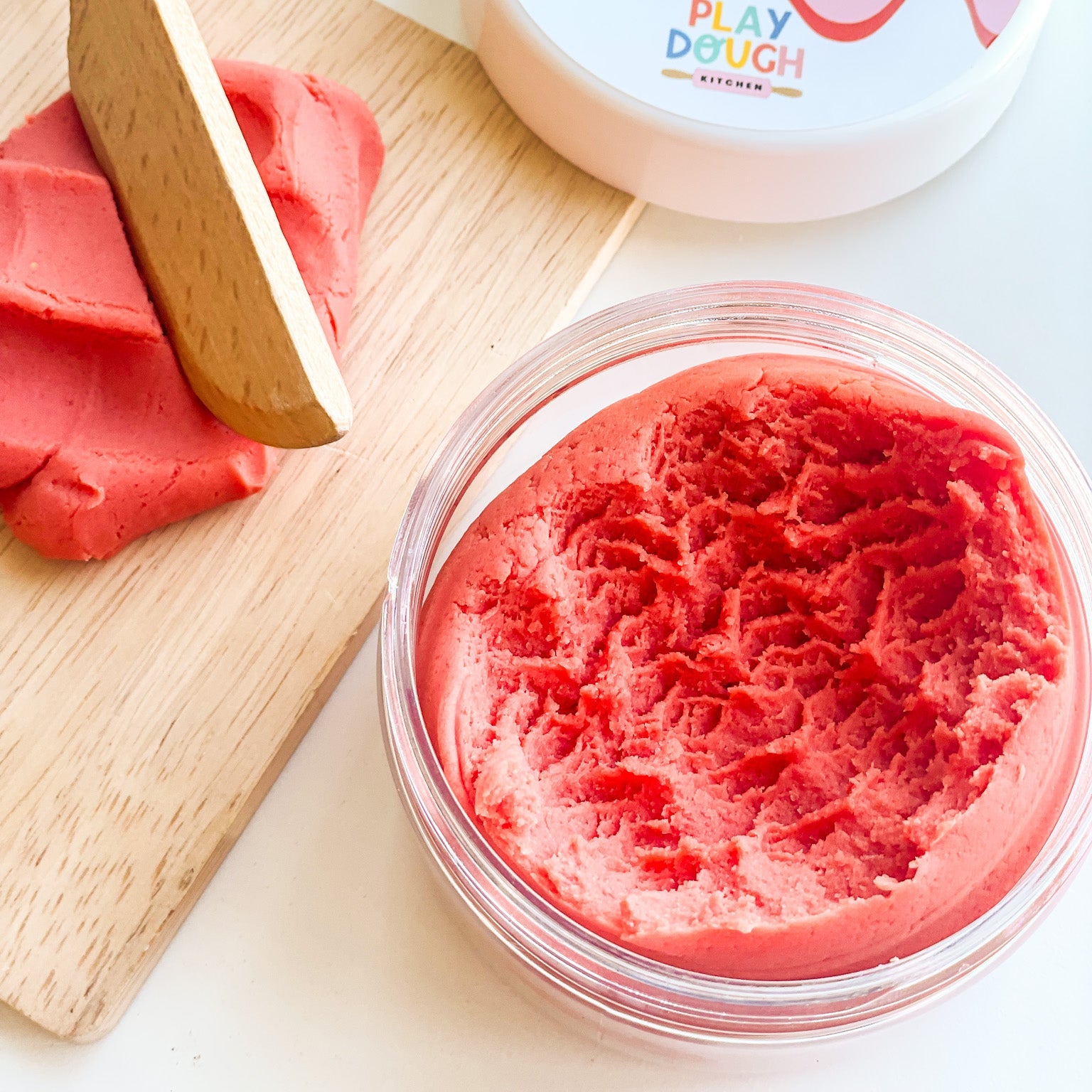 A jar of red cherry play dough on a white surface styled with a wooden chopping board and knife