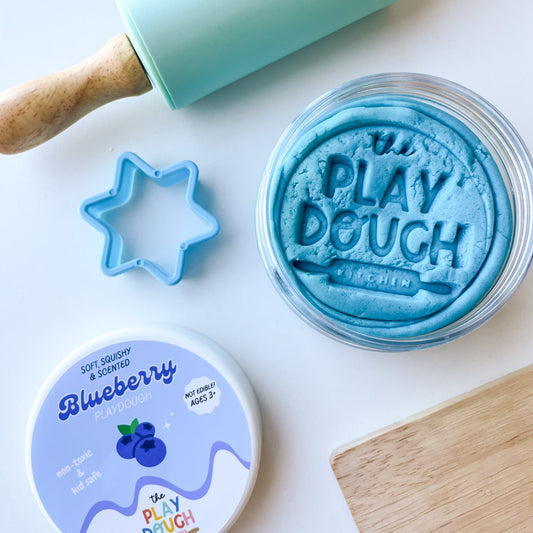 Blue play dough with a rolling pin, cookie cutter, and container on a white surface