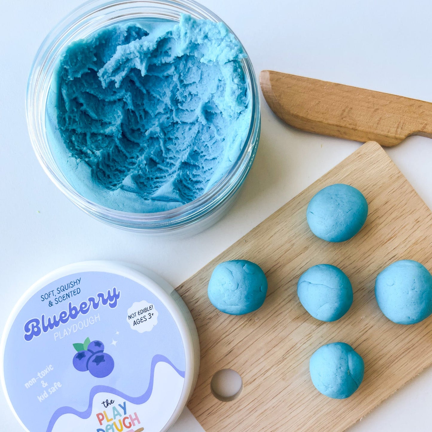 Blue play dough with a container, wooden board, and blueberry-themed packaging on a white background.