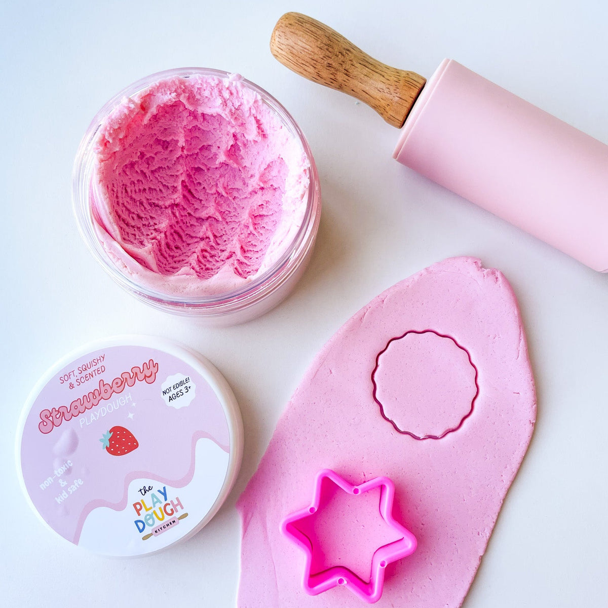 A jar of pink strawberry playdough on a white surface with cookie cutters and a rolling pin