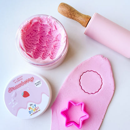 A jar of pink strawberry playdough on a white surface with cookie cutters and a rolling pin