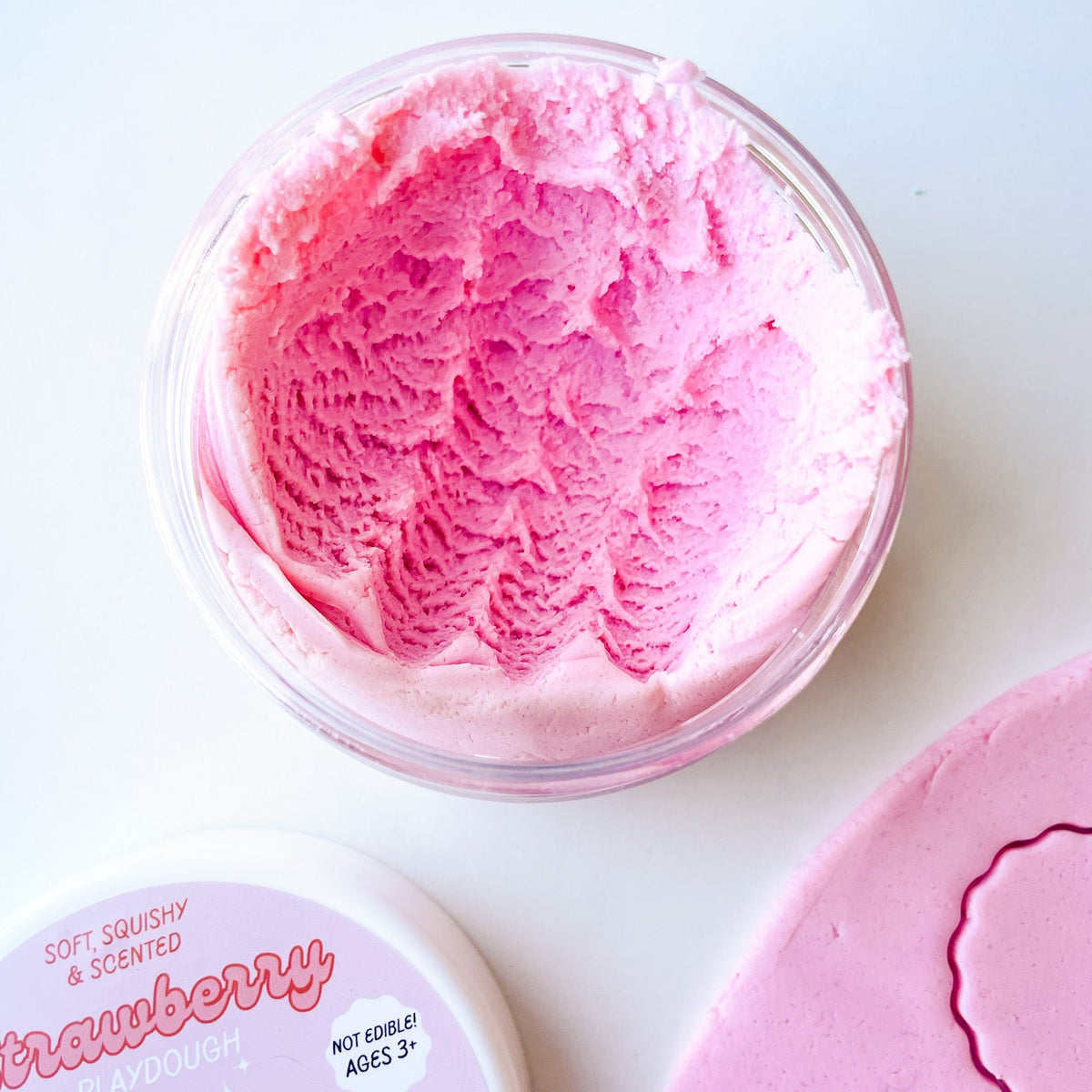 A clear jar of pink strawberry playdough on a white surface
